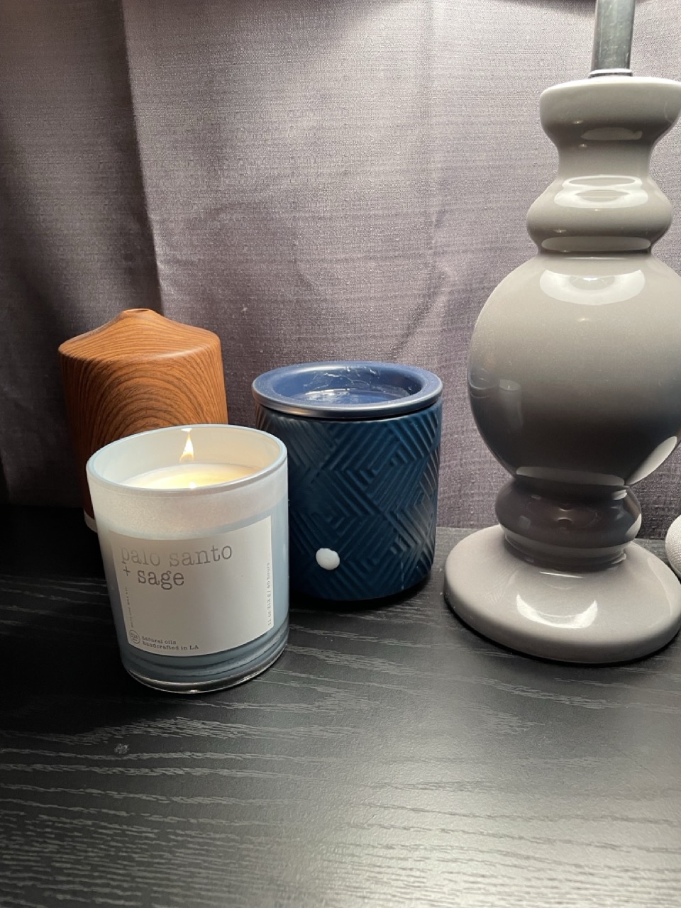 'Palo Santo + Sage' candle glowing beside a lamp and ceramic warmer.