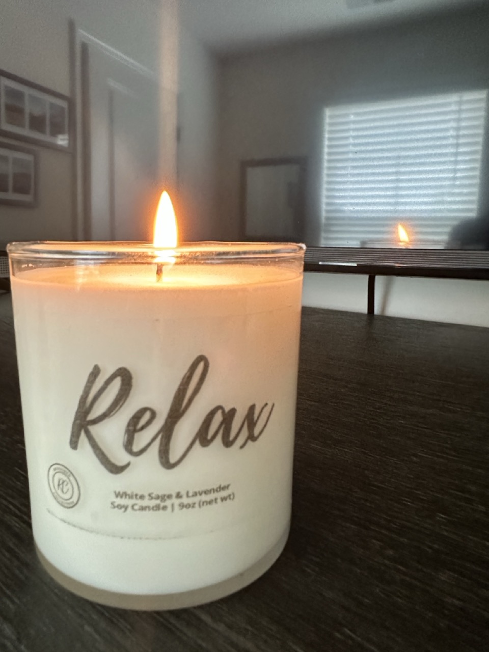 White candle labeled 'Relax' burning on a dark table with its reflection visible on a TV screen.